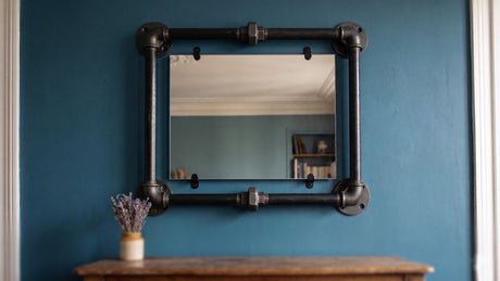 Cadre de miroir rectangulaire en tubes filetés et coudes fonte noire, fixé sur un mur bleu profond, fermé par deux raccords unions visibles au centre des barres horizontales — un exemple typique de cadre fermé en plomberie filetée décorative